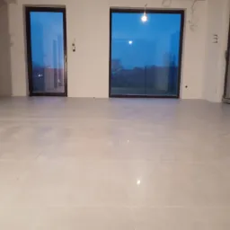 Jasna, świeżo położona podłoga z płytek w pustym pomieszczeniu z dużymi oknami i widocznymi przewodami elektrycznymi na ścianach.