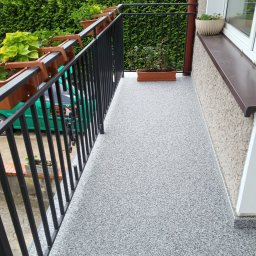 FIRMA REMONTOWO WYKOŃCZENIOWA PAWEŁ SARNA - Balkon z szarą, żywiczną posadzką, czarną balustradą z doniczkami i zielonymi roślinami na tle żywopłotu.