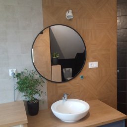 FIRMA REMONTOWO WYKOŃCZENIOWA PAWEŁ SARNA - Nowoczesna łazienka z umywalką nablatową, okrągłym lustrem z czarną ramą, drewnopodobnymi płytkami ułożonymi w geometryczny wzór na ścianie i rośliną doniczkową obok.