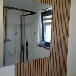 Nowoczesna łazienka z kabiną prysznicową w kolorze czarnym, widoczna w lustrze, obok ściana z drewnianych paneli dekoracyjnych.