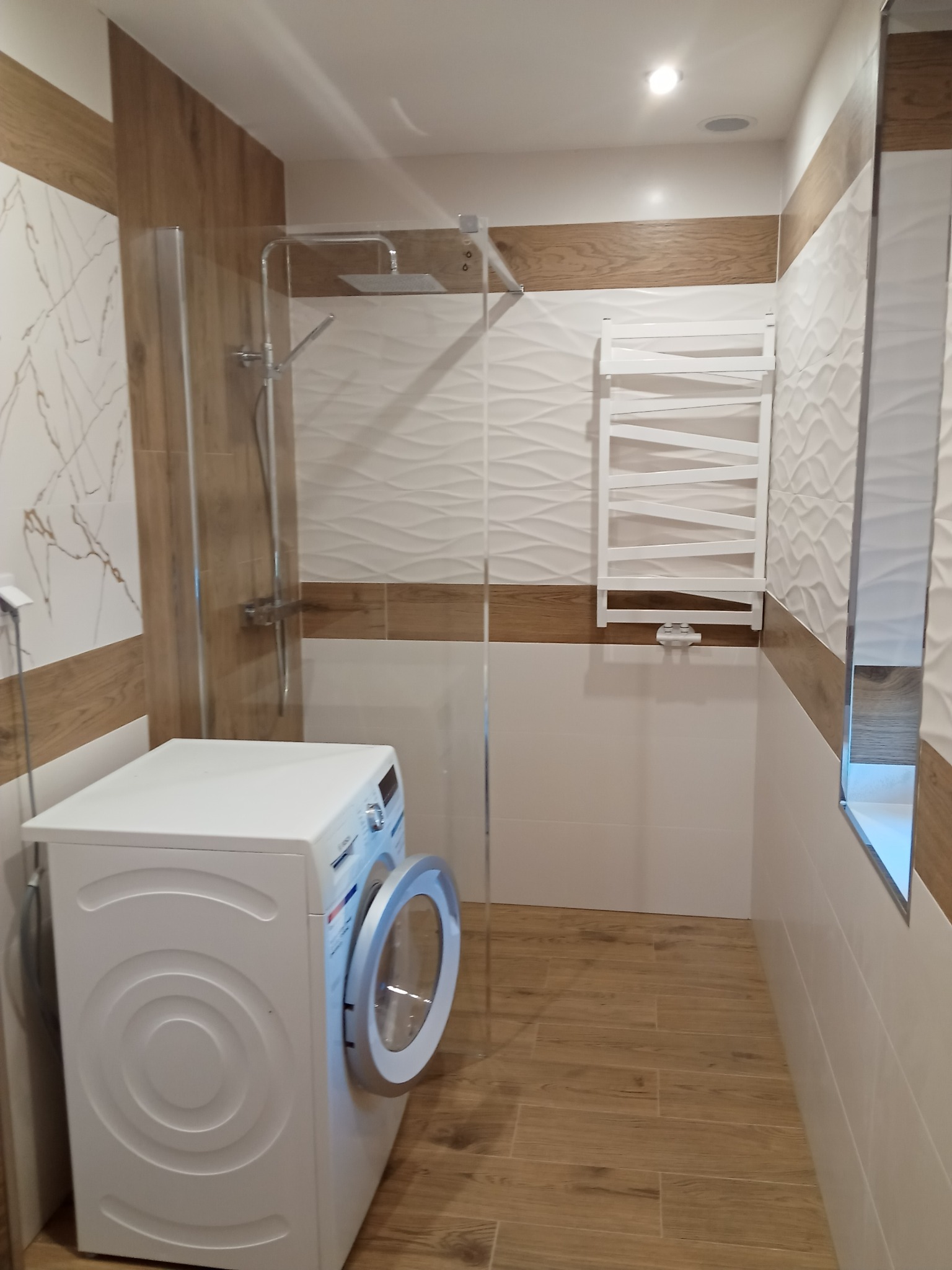 Nowoczesna łazienka z kabiną prysznicową typu walk-in, pralką z otwartymi drzwiczkami, białymi płytkami z subtelnym wzorem fal i drewnopodobnymi akcentami.