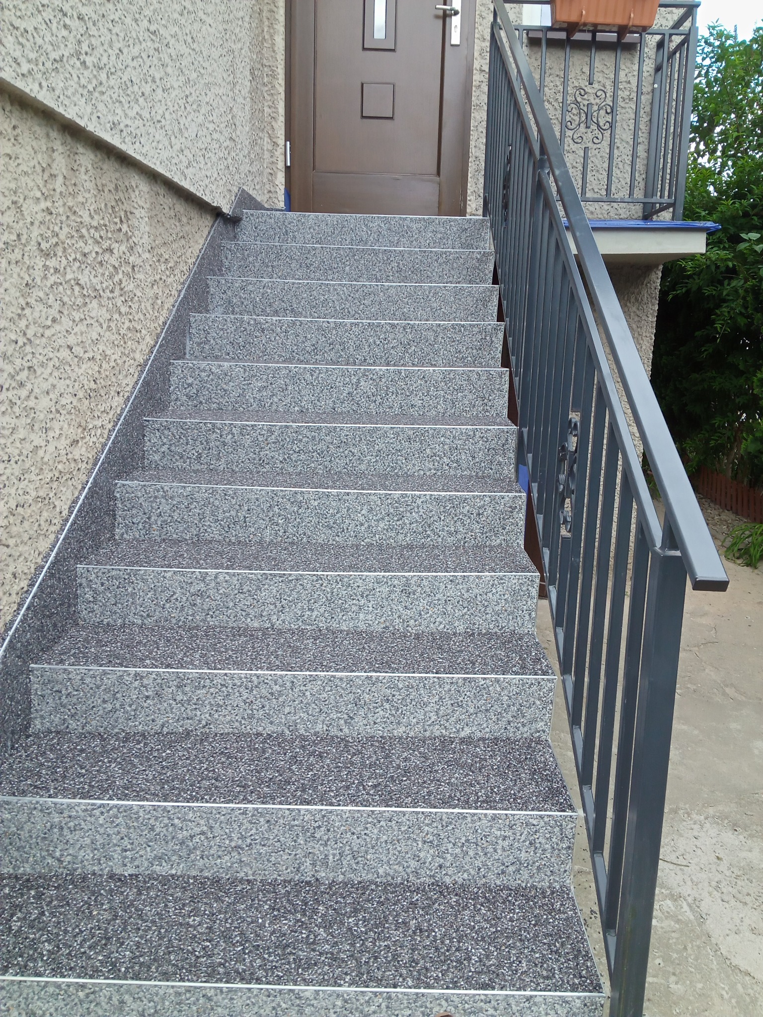 Zewnętrzne schody wejściowe wyłożone szarą, drobnoziarnistą mozaiką z metalowymi listwami antypoślizgowymi, prowadzące do brązowych drzwi wejściowych z metalową balustradą po prawej stronie.