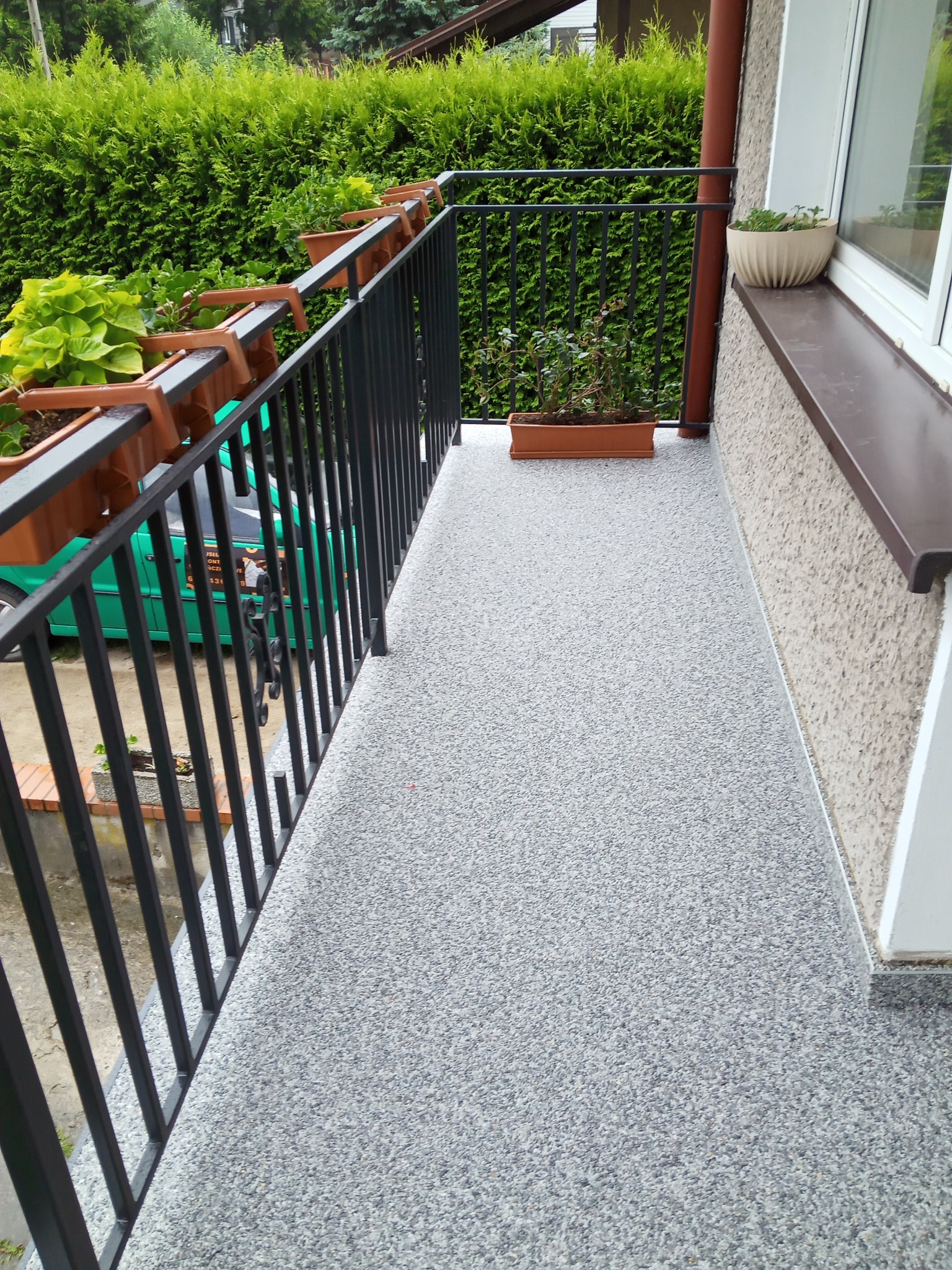 Balkon z szarą, żywiczną posadzką, czarną balustradą z doniczkami i zielonymi roślinami na tle żywopłotu.
