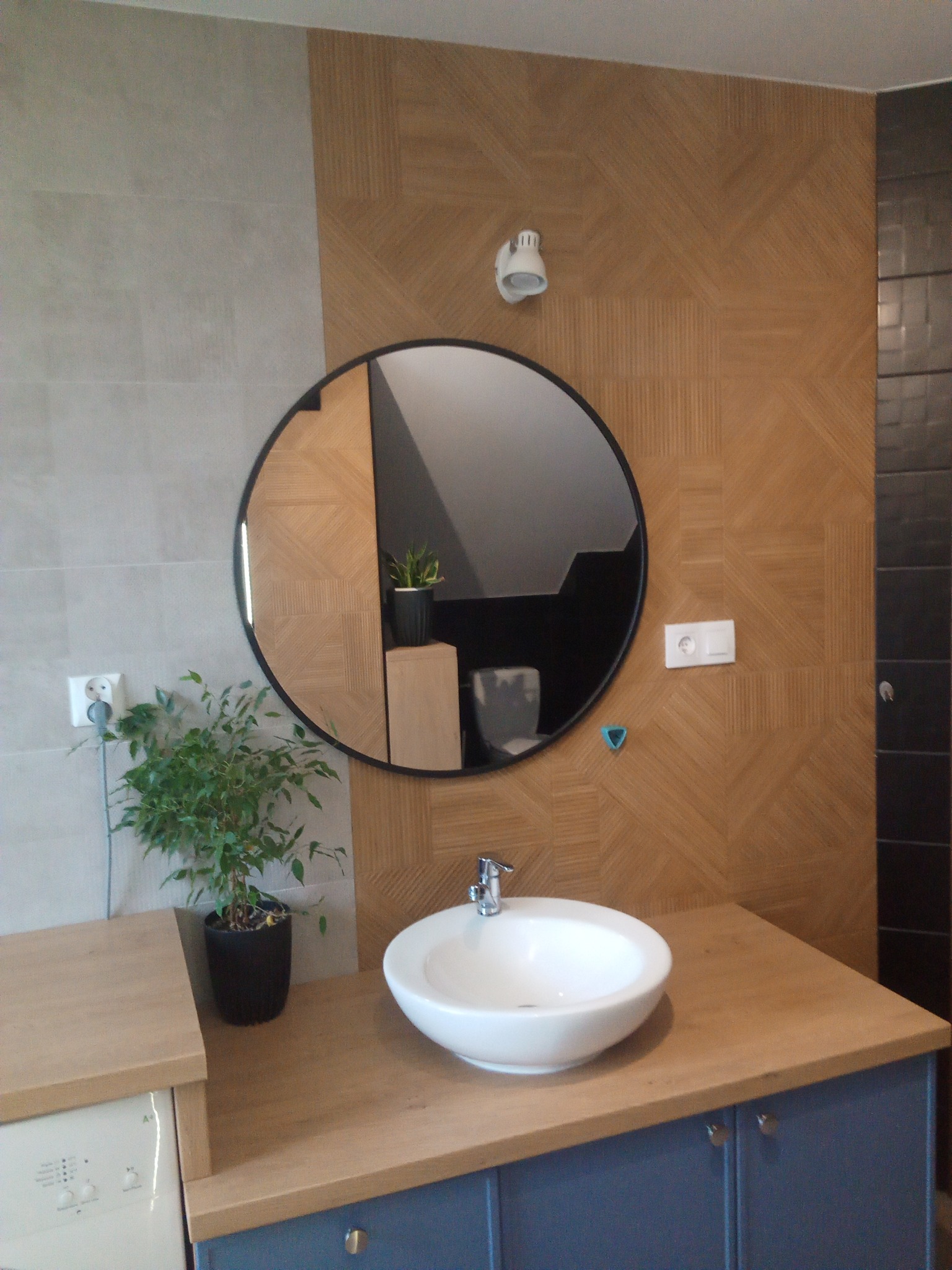 Nowoczesna łazienka z umywalką nablatową, okrągłym lustrem z czarną ramą, drewnopodobnymi płytkami ułożonymi w geometryczny wzór na ścianie i rośliną doniczkową obok.