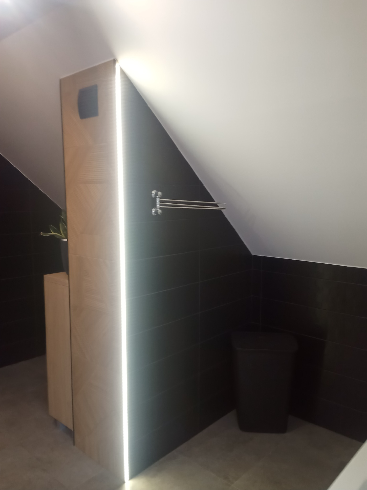 Nowoczesna łazienka na poddaszu z czarnymi płytkami, oświetleniem LED wzdłuż zabudowy meblowej z drewnopodobnym dekorem oraz grzejnikiem drabinkowym i koszem na śmieci.