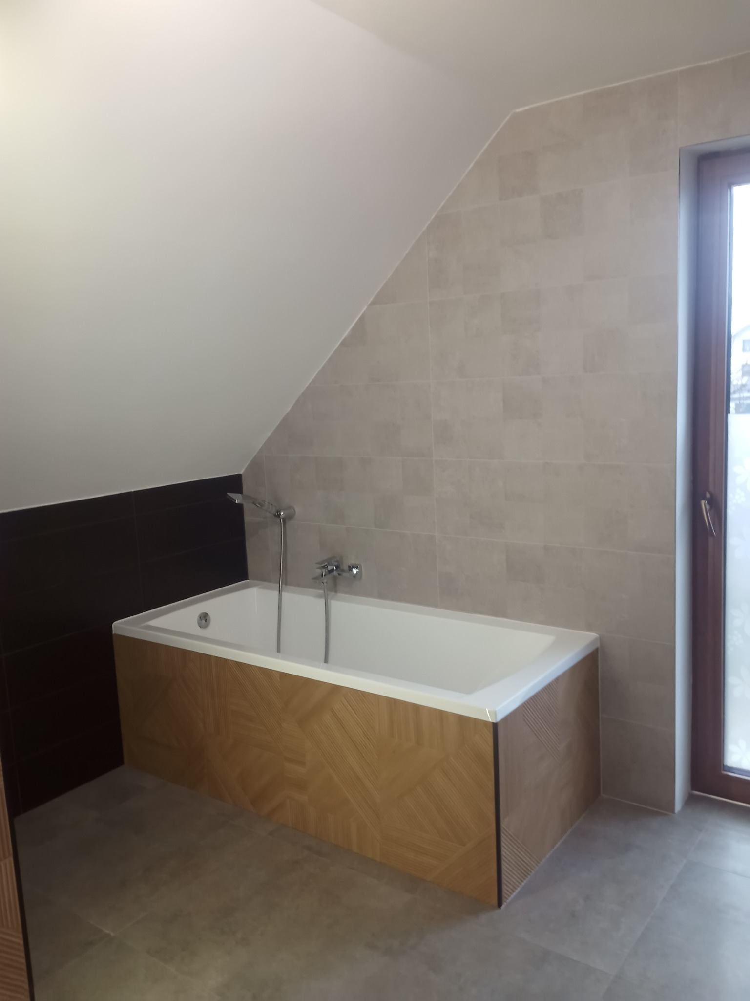 Nowoczesna łazienka na poddaszu z wanną obudowaną drewnopodobną mozaiką, beżowe płytki na ścianach, brązowe płytki przy wannie, okno z dekoracyjną folią.