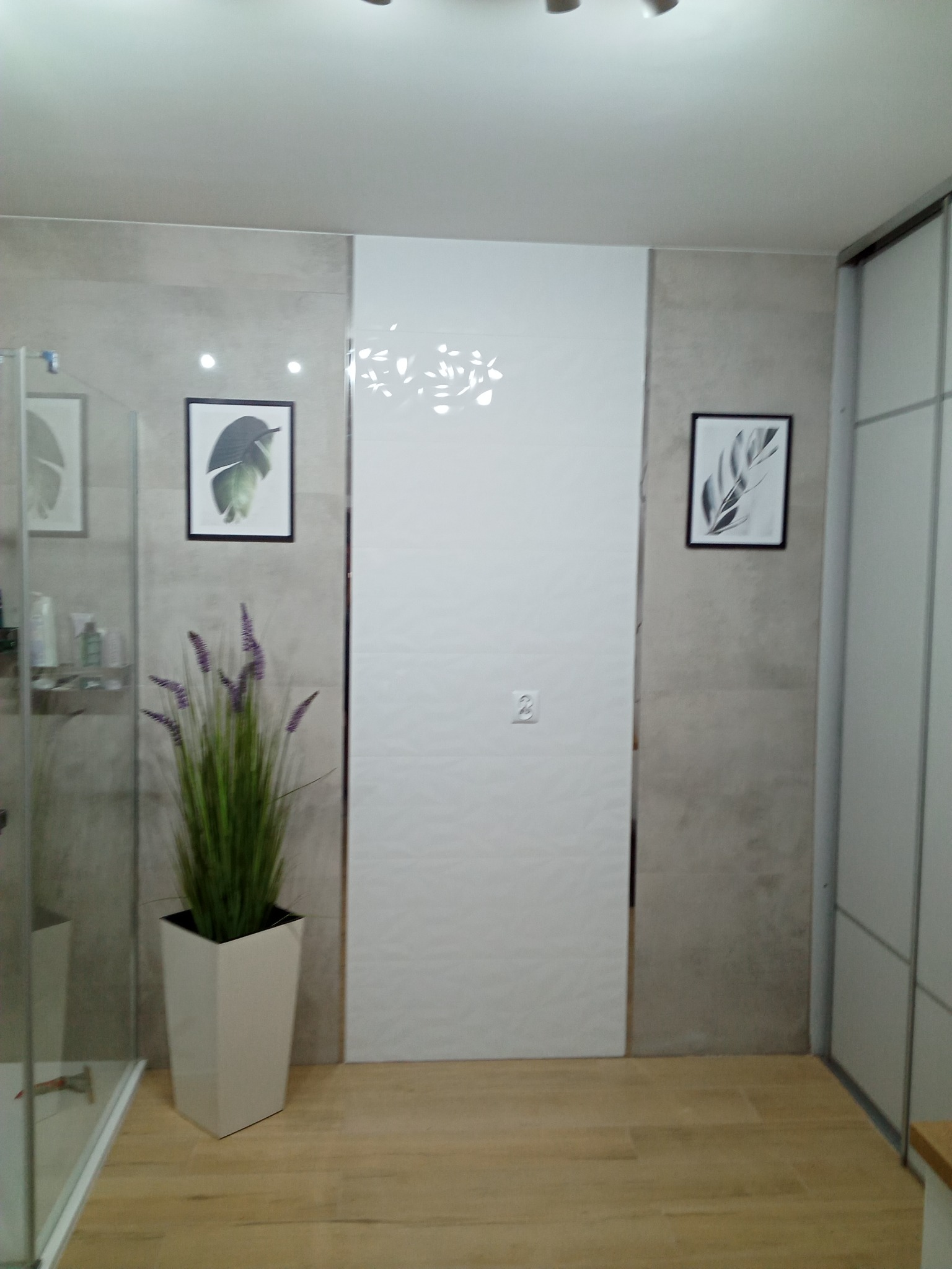 Nowoczesna łazienka z białymi panelami ściennymi o subtelnym wzorze, drewnopodobną podłogą, minimalistycznymi obrazami w czarnych ramkach i wysoką donicą z fioletowymi kwiatami obok kabiny...