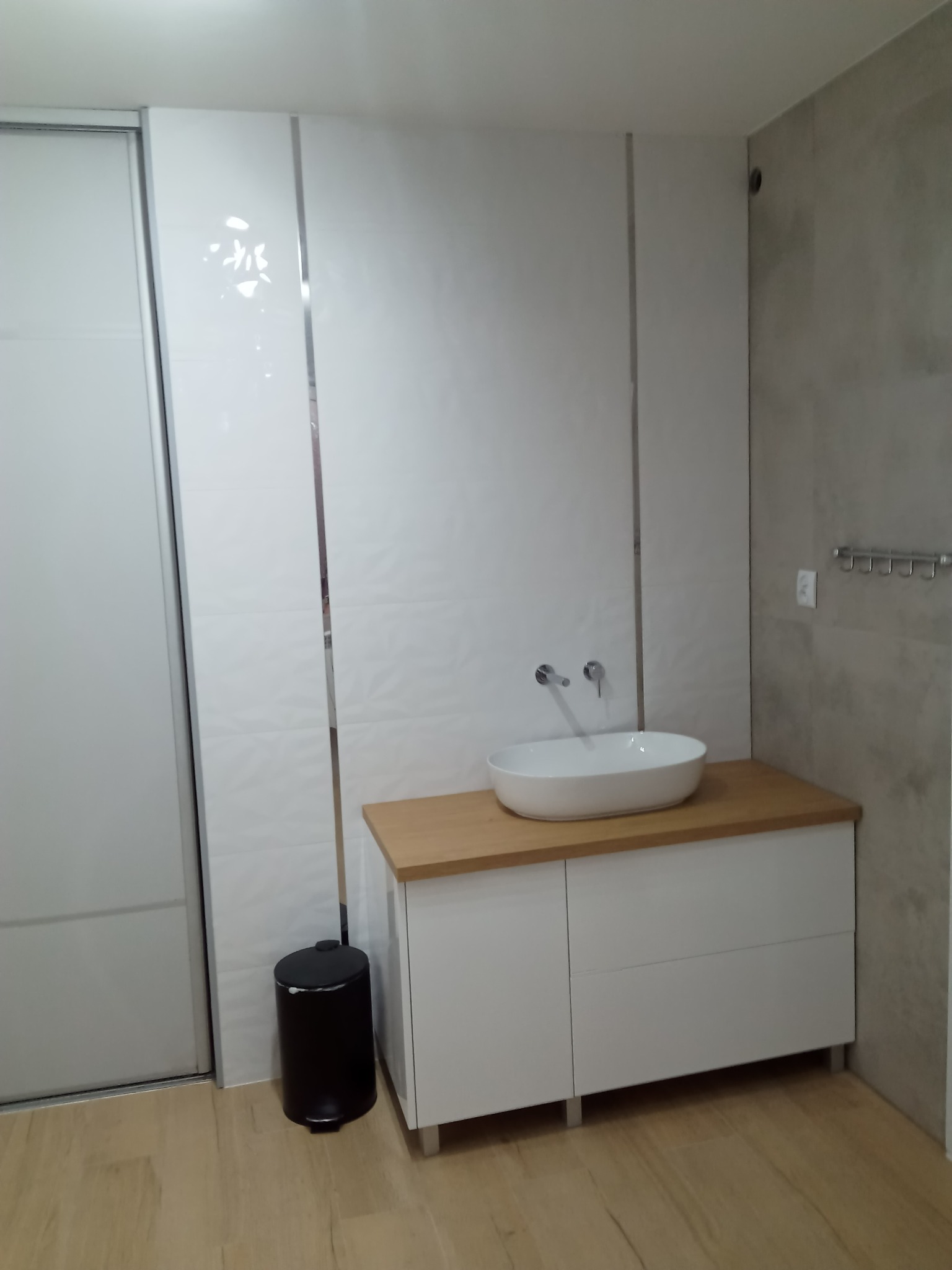 Nowoczesna łazienka po remoncie: biała umywalka nablatowa na szafce z drewnianym blatem, białe płytki z subtelnym wzorem i srebrnymi listwami, szara ściana z wieszakami, podłoga z paneli...