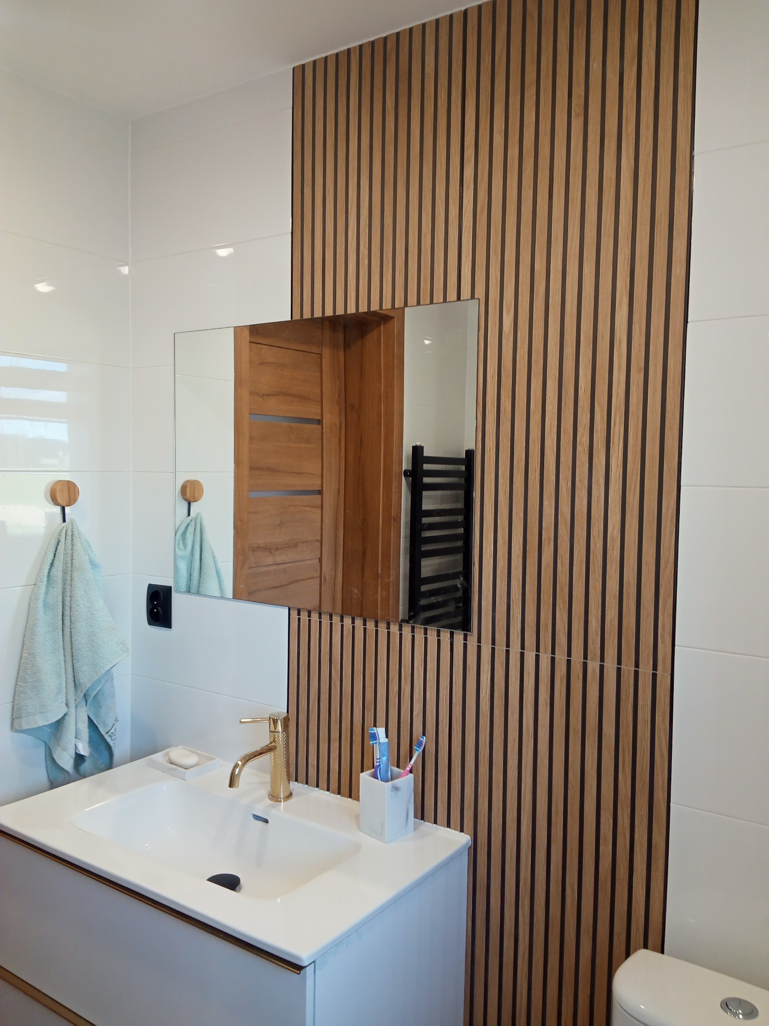 Nowoczesna łazienka z drewnianymi lamelami na ścianie, białą umywalką z złotą armaturą i lustrem odbijającym drewniane drzwi.