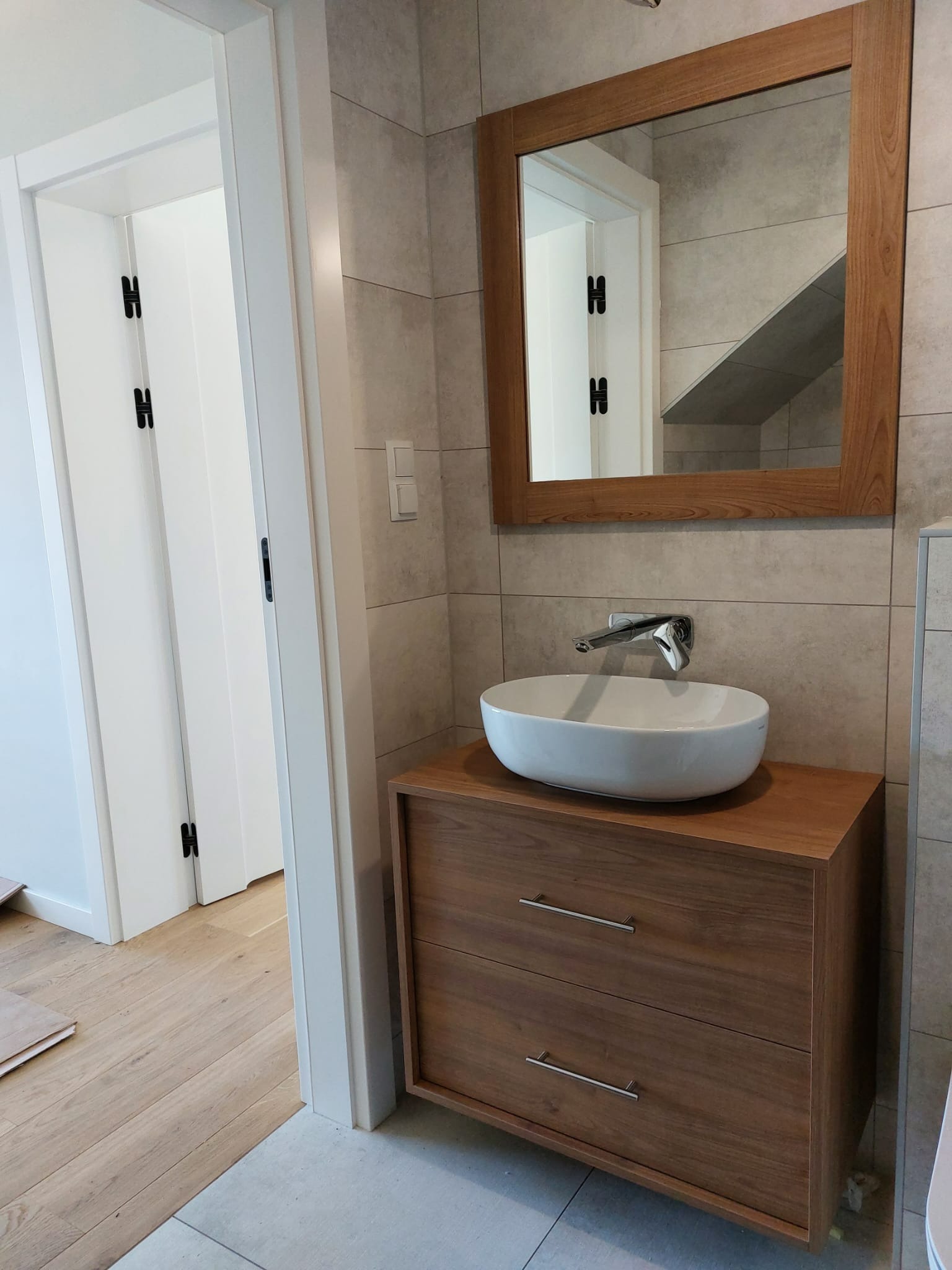 Nowoczesna łazienka z drewnianą szafką pod umywalkę nablatową, lustrem w drewnianej ramie i szarą płytką na ścianach; widoczny fragment podłogi z drewnianych paneli oraz drzwi z czarnymi zawiasami.