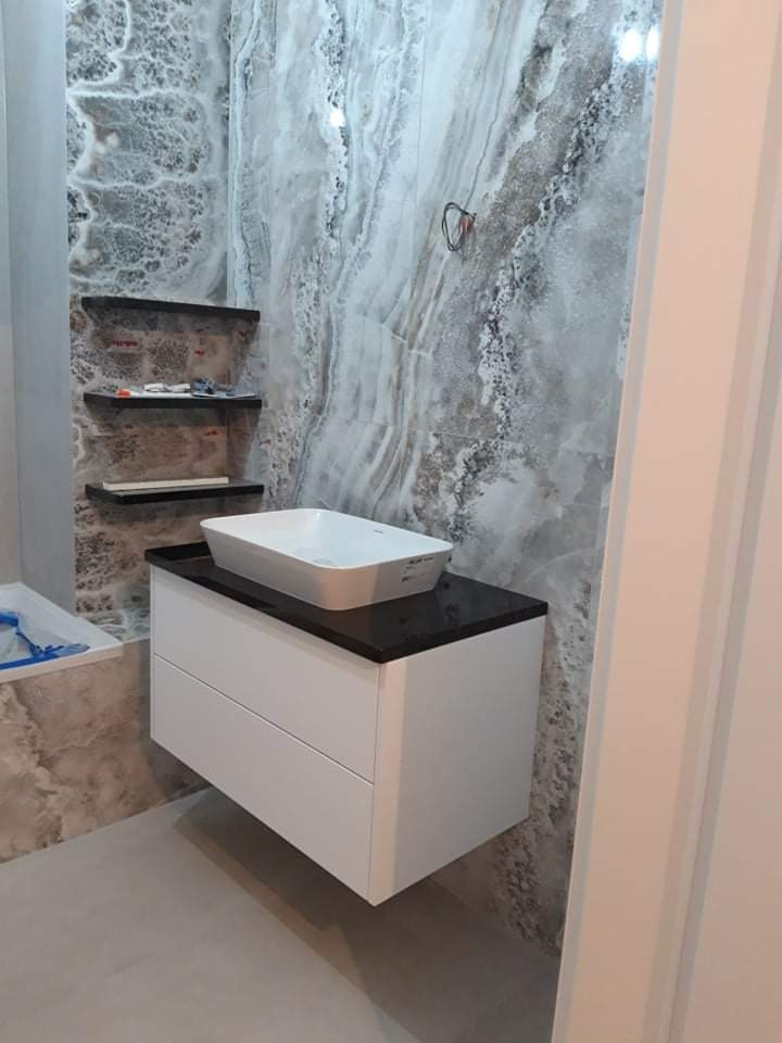 Łazienka w trakcie wykańczania: biała szafka pod umywalkę z czarnym blatem, umywalka nablatowa, marmurowe płytki na ścianach, widoczne półki i niedokończone instalacje.