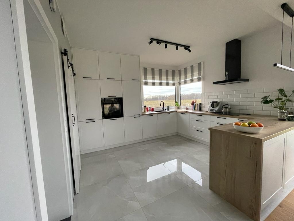 Nowoczesna kuchnia z białymi szafkami, czarnymi uchwytami, drewnianym blatem wyspy kuchennej i szarymi płytkami podłogowymi.