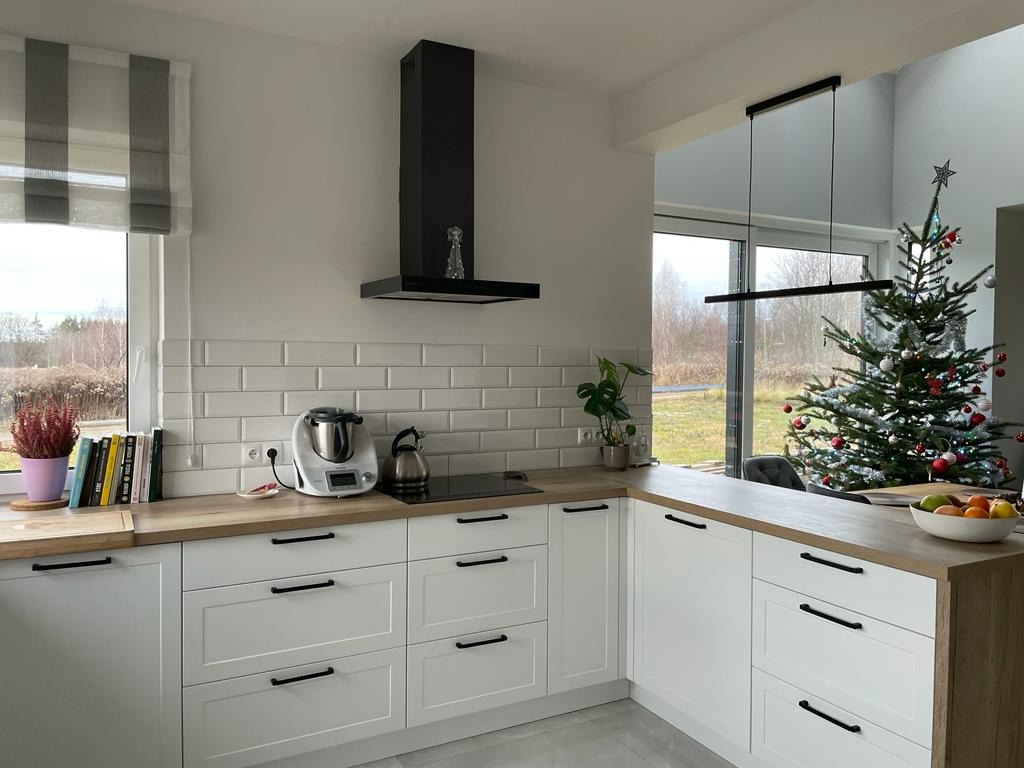 Jasna kuchnia z białymi szafkami i drewnianym blatem, czarnym okapem i minimalistycznym oświetleniem, z widocznym fragmentem choinki w tle.