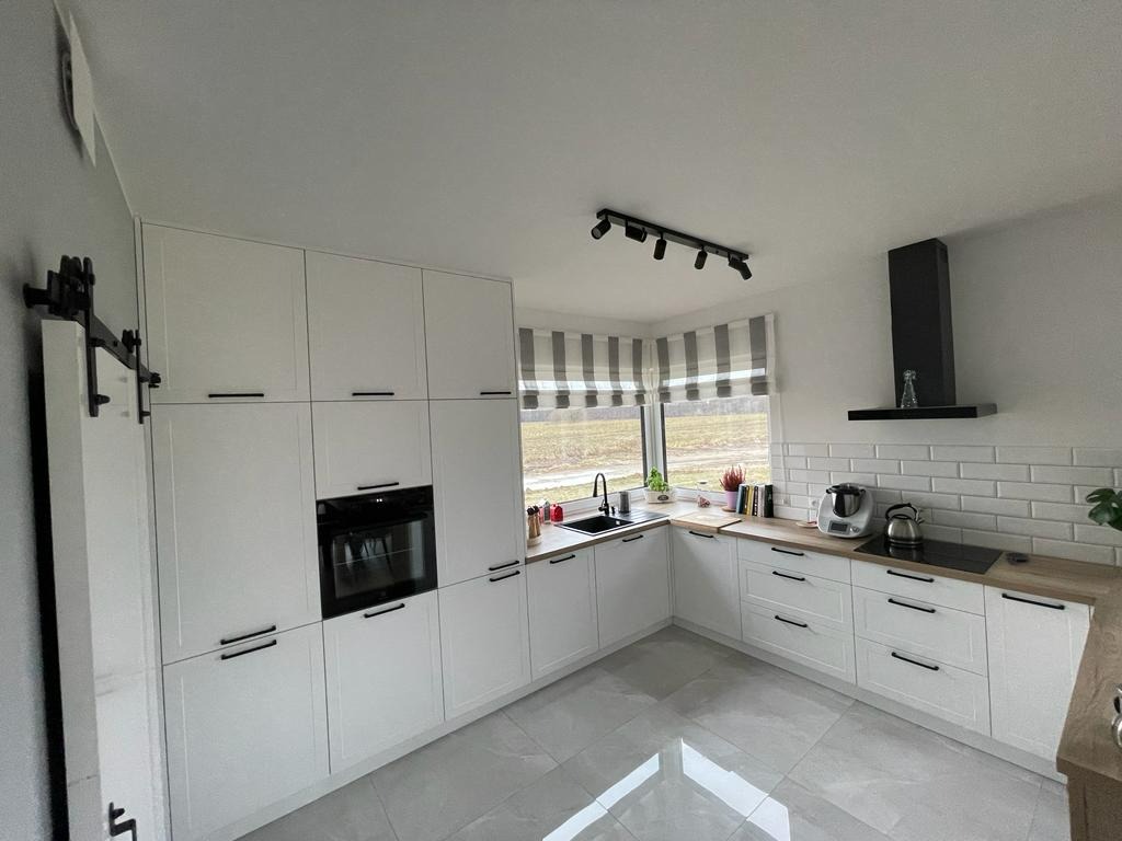 Jasna kuchnia z białymi szafkami, drewnianymi blatami i szarymi płytkami na podłodze, widok na okno z roletami w paski.