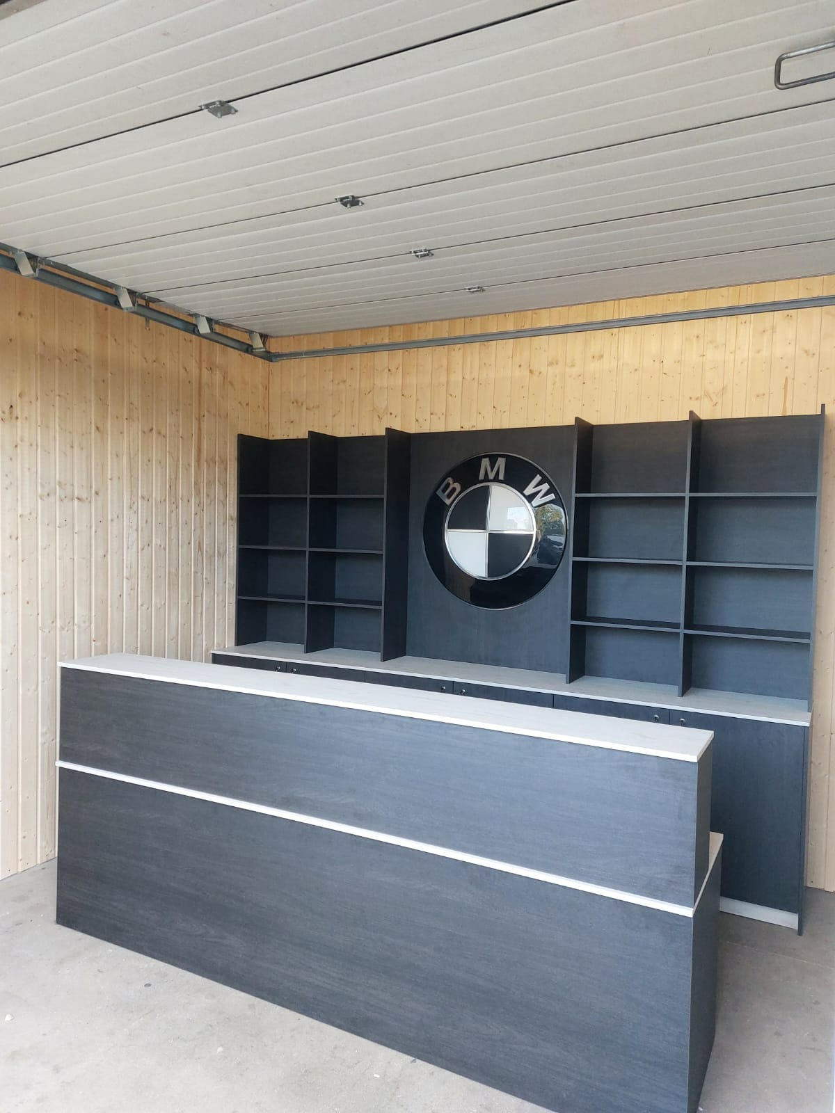 Recepcyjna lada z szarego drewna z białymi akcentami, za nią regały i szafki z logo BMW w okrągłej obudowie, drewniana boazeria na ścianie, jasne pomieszczenie.