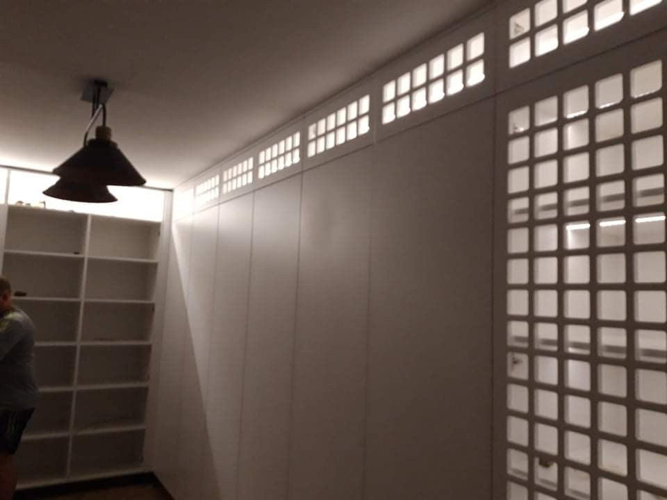 Biała zabudowa meblowa od podłogi do sufitu z ażurowymi elementami wentylacyjnymi w górnej części i na skrzydle drzwiowym, oświetlona wiszącą lampą z dwoma kloszami.