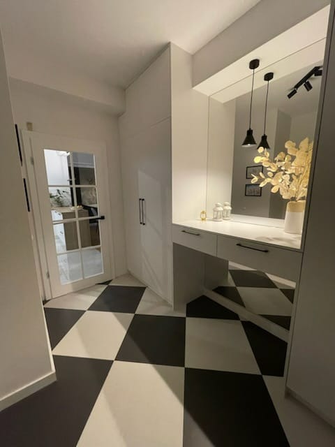 Wnętrze przedpokoju z zabudowaną szafą, toaletką z lustrem, czarnymi lampami wiszącymi i podłogą w szachownicę.