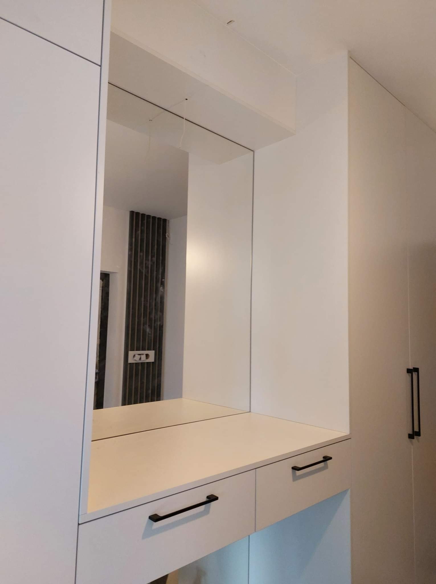 Biała zabudowa meblowa z lustrem, widoczne czarne, minimalistyczne uchwyty szuflad i szafek, w lustrze odbija się fragment ściany z dekoracyjnymi panelami.