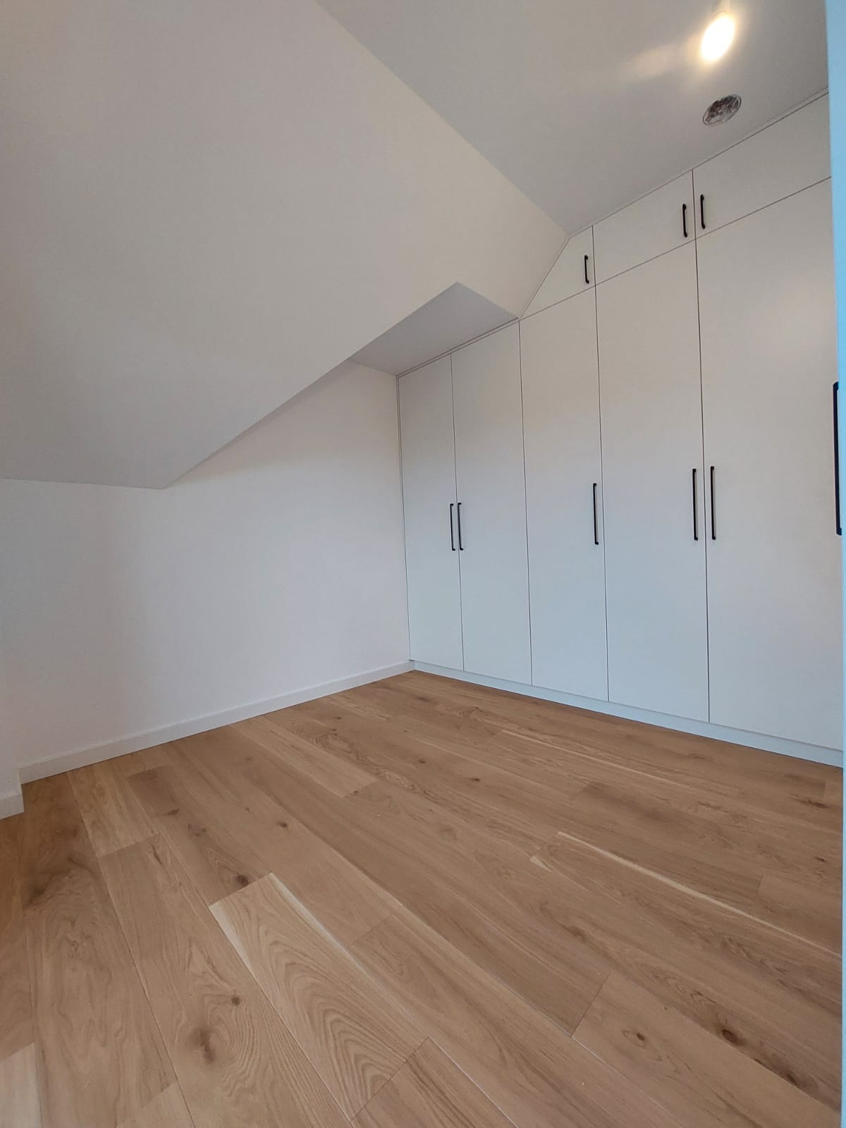 Wnętrze poddasza ze skośnym sufitem, białe szafy w zabudowie z czarnymi, długimi uchwytami, drewniana podłoga z jasnych desek.