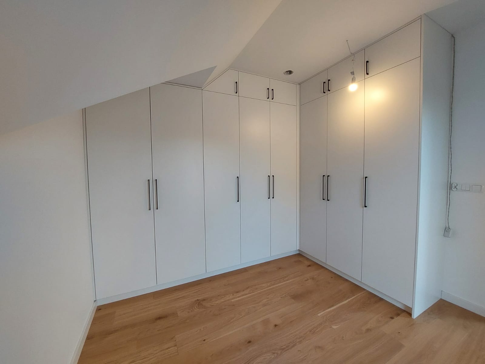 Biała szafa wnękowa z czarnymi, długimi uchwytami, dopasowana do skosu poddasza, z drewnianą podłogą