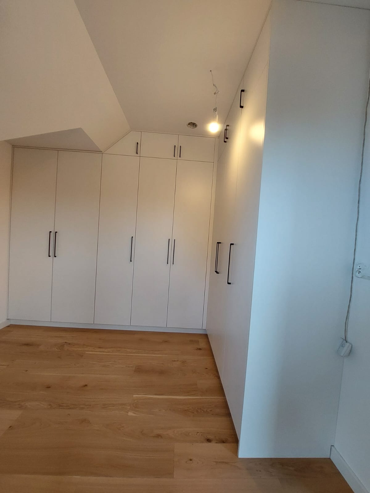 Biała zabudowa szaf wnękowych z czarnymi uchwytami, dopasowana do skosu dachu, na tle drewnianej podłogi.