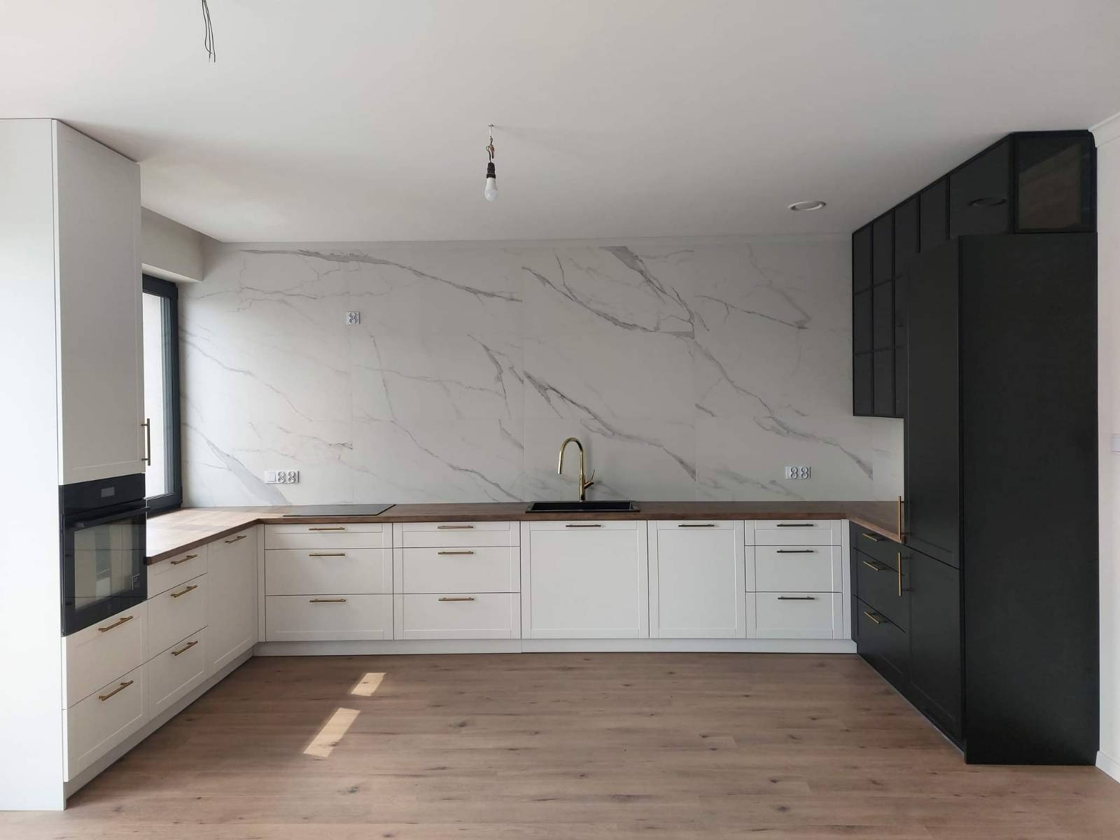 Nowoczesna kuchnia z białymi szafkami dolnymi i czarnymi górnymi, drewnianym blatem i marmurową ścianą, złote uchwyty, surowa żarówka zwisająca z sufitu.