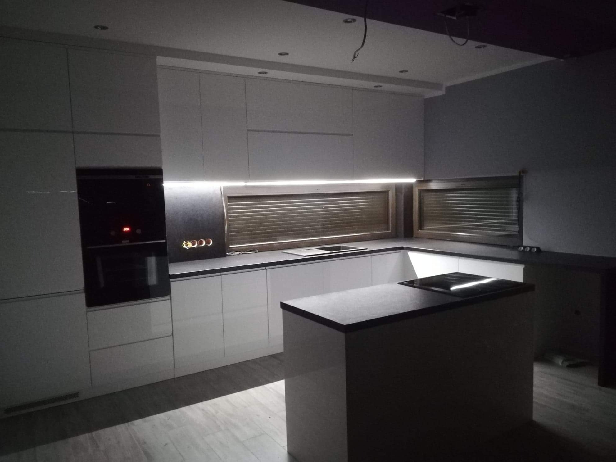 Nowoczesna, biała kuchnia z wyspą i ciemnym blatem, oświetlenie podszafkowe LED, widoczne gniazdka elektryczne nad blatem, częściowo zaciemnione okno.