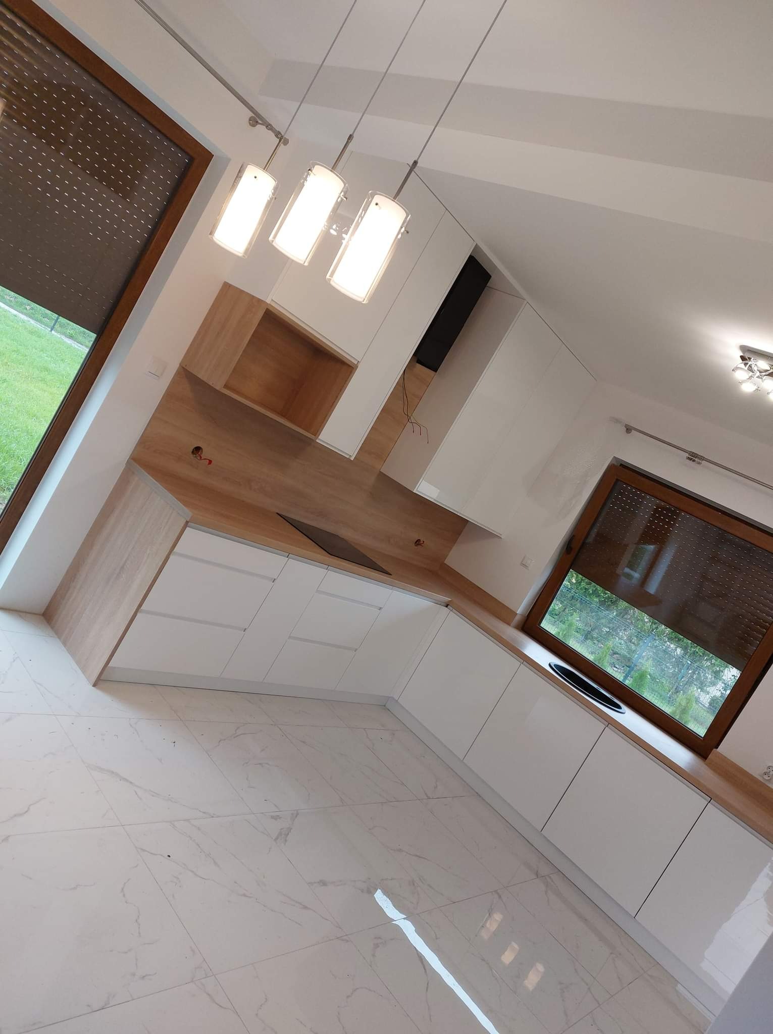 Nowoczesna kuchnia z białymi frontami szafek i drewnianym blatem roboczym, widok z perspektywy podłogi, z trzema wiszącymi lampami i dużym oknem z roletami zewnętrznymi.