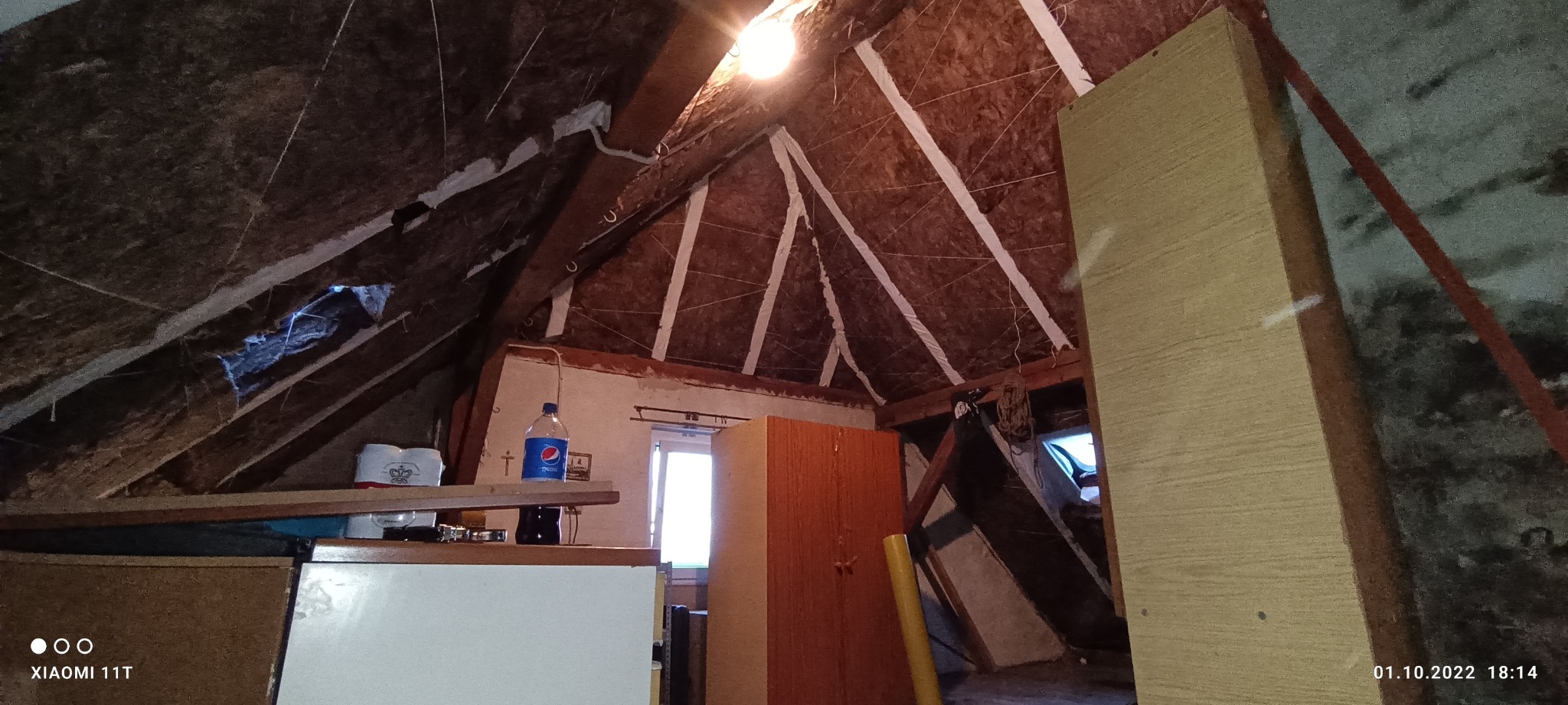 Poddasze z widoczną izolacją termiczną na skośnym dachu, częściowo wykończone, z widocznym oknem, meblami i butelką napoju na blacie.