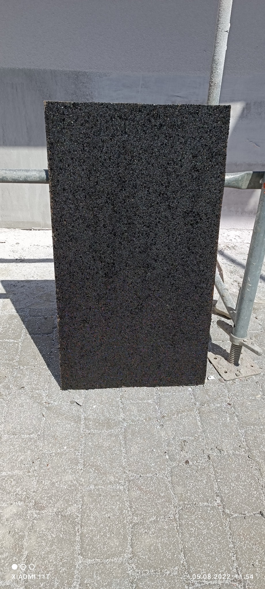 Ciemna płyta izolacyjna z widoczną strukturą, oparta o chodnik z kostki brukowej, obok rusztowania.