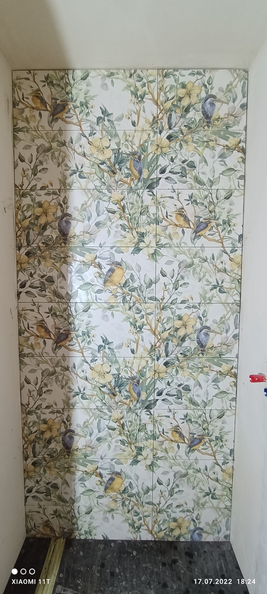 Ułożone płytki ceramiczne z motywem roślinnym i ptaków na ścianie w łazience, widoczne fugi między płytkami.