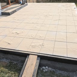 Usługi Ogólnobudowlane Sławomir Kmiecik - Jasne płyty tarasowe w trakcie układania, posypane piaskiem, widoczne ślady butów, drewniana deska pomocnicza na trawie, fragment budynku w tle.