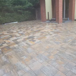 Nowa, wielobarwna kostka brukowa ułożona na posesji, widok z góry. W tle budynek z ceglanymi kolumnami i zielone drzewa. Kostka w odcieniach szarości, beżu i brązu.