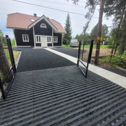 Usługi Ogólnobudowlane Sławomir Kmiecik - Czarna, ażurowa nawierzchnia podjazdu z jasnymi pasami, prowadząca do ciemnego domu z czerwoną dachówką. Otwarta brama w tle, drzewa i tabliczka ostrzegawcza.