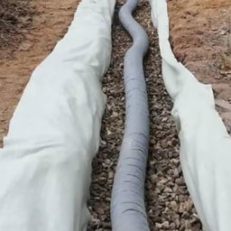 Usługi Ogólnobudowlane Sławomir Kmiecik - Wykop z rurą drenażową, obsypany żwirem i wyłożony białą geowłókniną. W tle widać ziemię i drzewa. Na skraju kadru taczka.