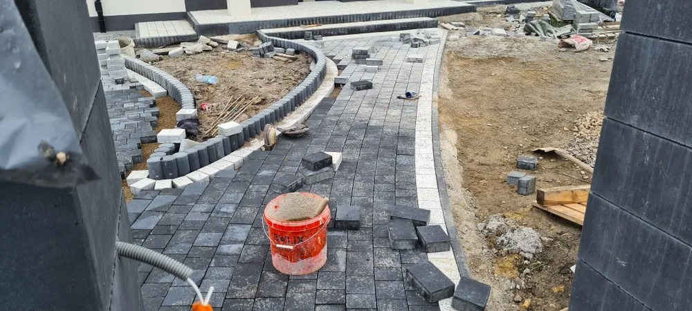 Plac budowy z częściowo ułożoną, szarą kostką brukową. Widoczny czerwony pojemnik z zaprawą, obrzeża z kostki, przygotowany teren pod dalsze prace i narzędzia budowlane.