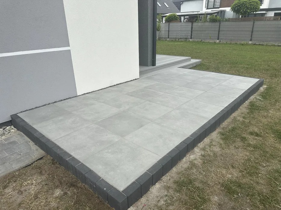 Nowoczesny taras wyłożony jasnoszarą płytką kwadratową, obrzeża z ciemnoszarej kostki, przylegający do budynku z elewacją w odcieniach szarości i bieli, trawnik w tle.