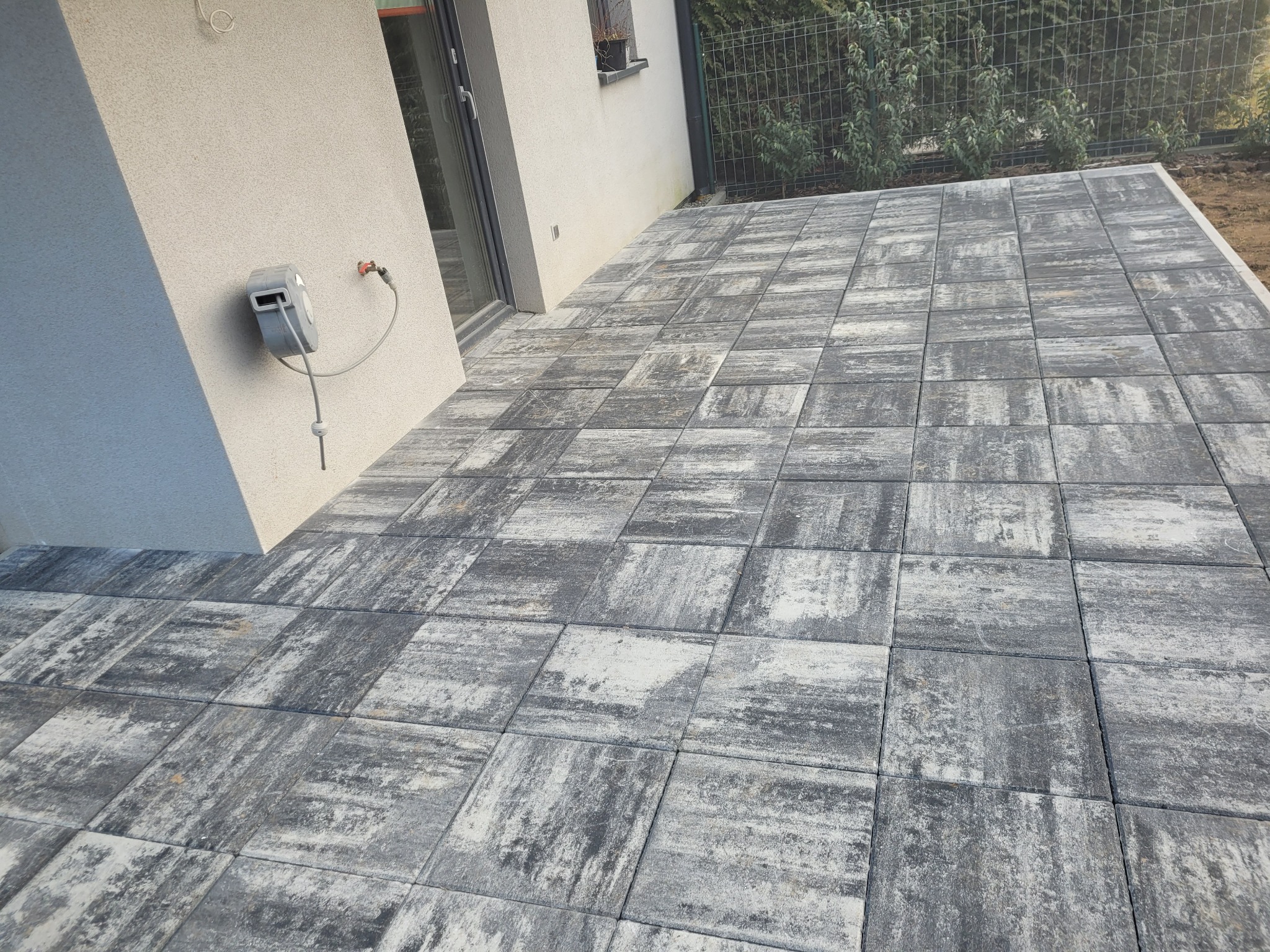 Nowoczesny taras wyłożony szaro-białą kostką brukową o kwadratowym kształcie, przylegający do ściany budynku z zamontowanym zwijadłem do węża ogrodowego.