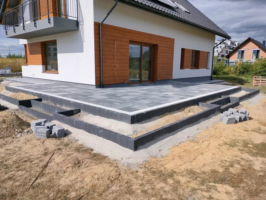 Tarasy betonowe w trakcie budowy z obrzeżami z kostki, widoczne niedokończone krawędzie i zapas kostki, dom jednorodzinny w tle.