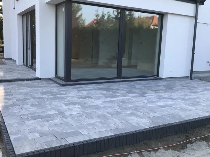 Nowoczesny taras wyłożony szarą kostką brukową w kształcie prostokątów, obrzeża wykończone ciemniejszą kostką pionowo, duże okna przesuwne w tle, biała elewacja budynku.