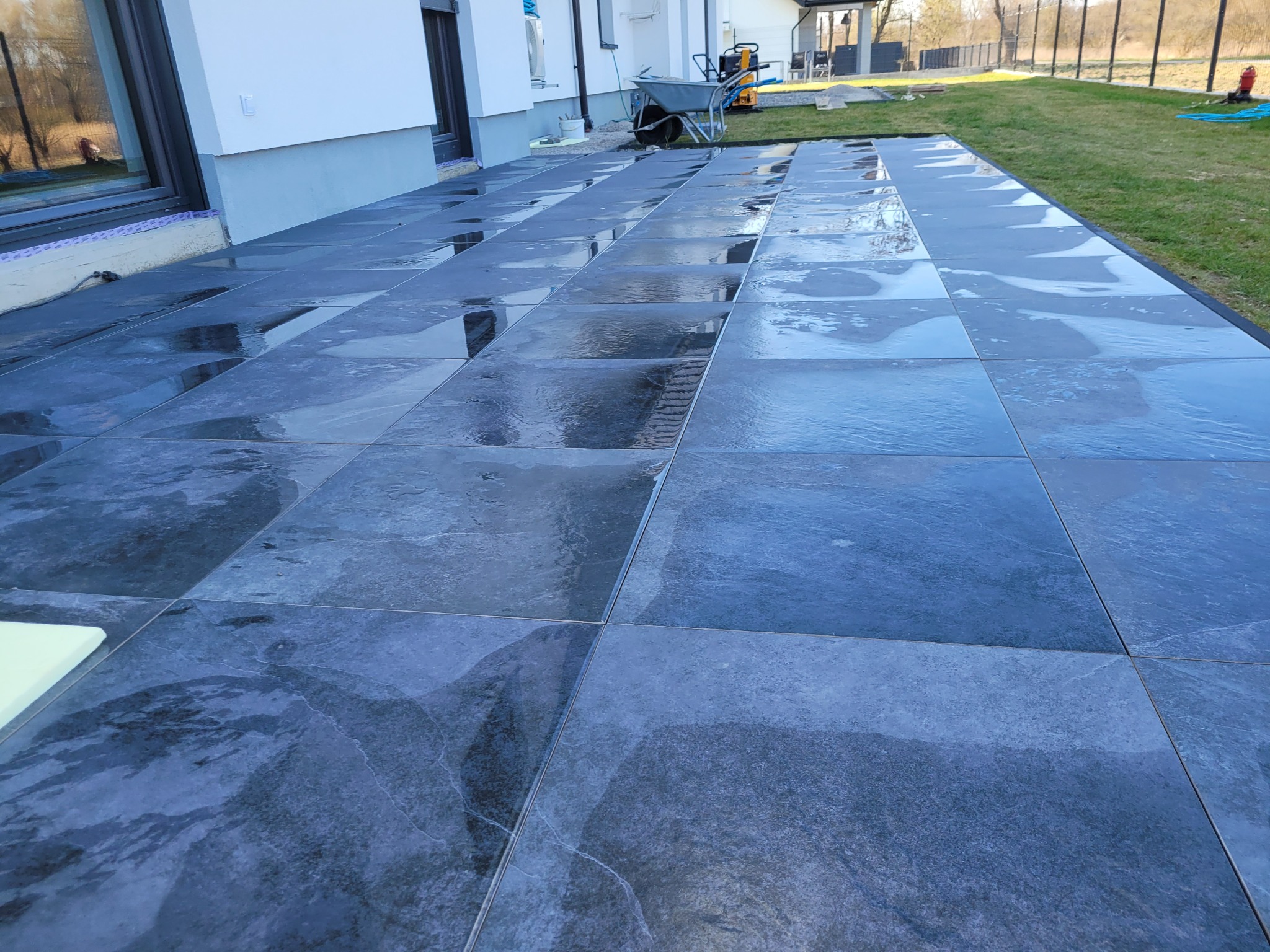 Świeżo ułożony, mokry taras z ciemnych, kwadratowych płyt kamiennych; w tle widoczny dom z dużym oknem przesuwnym, taczka i fragment trawnika.