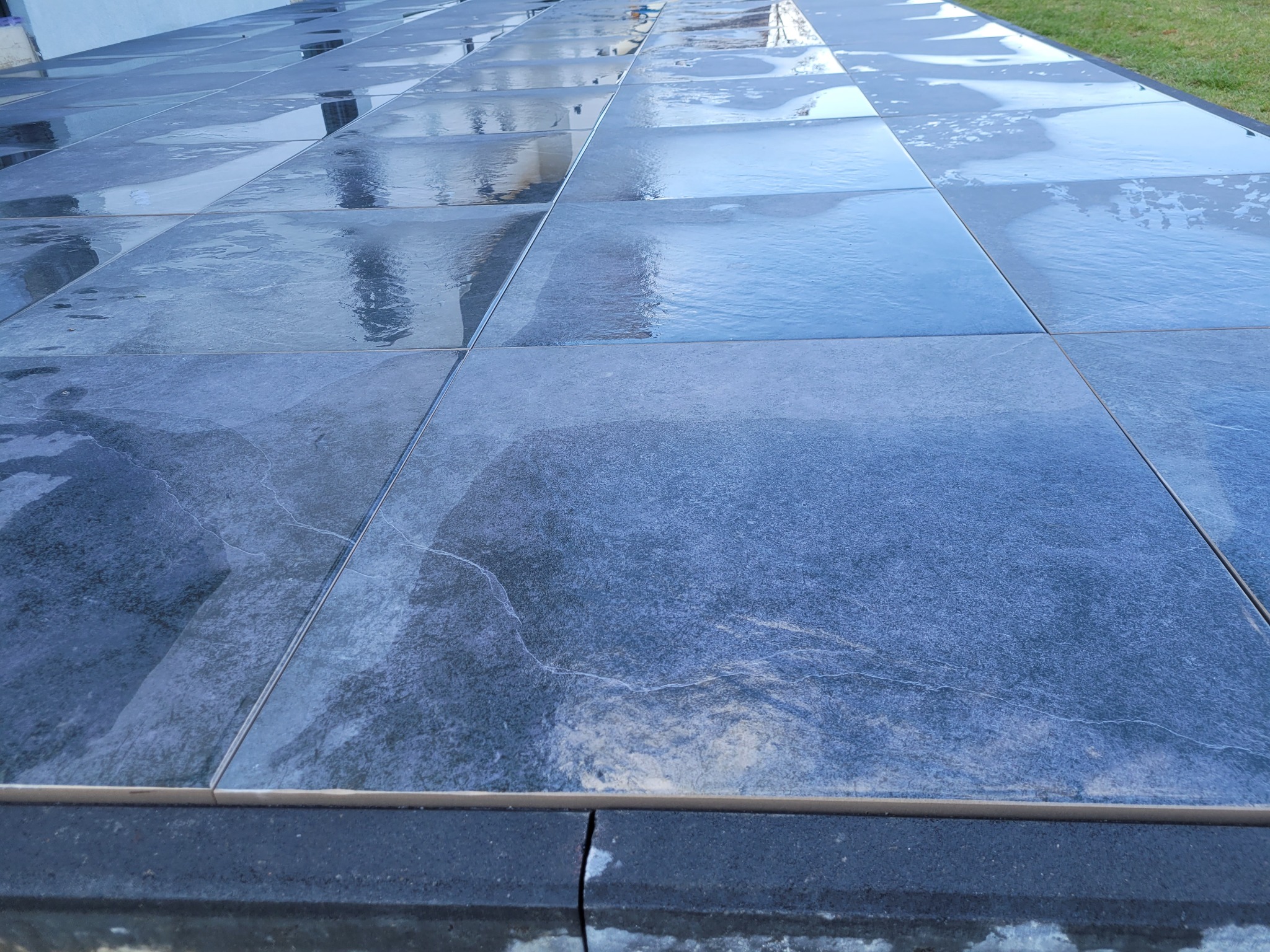 Świeżo ułożony taras z grafitowych płyt kamiennych, z widocznymi refleksami wody na powierzchni i zielenią trawnika w tle.