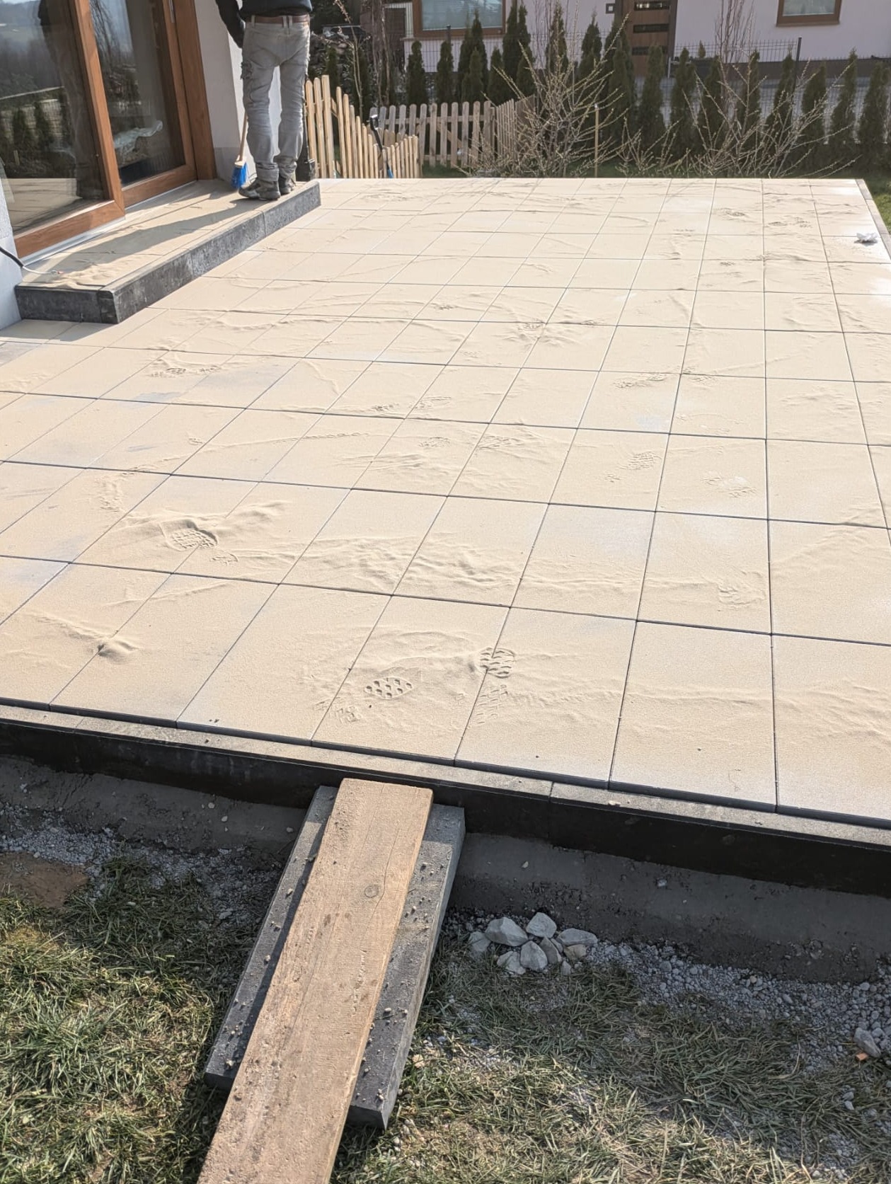 Jasne płyty tarasowe w trakcie układania, posypane piaskiem, widoczne ślady butów, drewniana deska pomocnicza na trawie, fragment budynku w tle.
