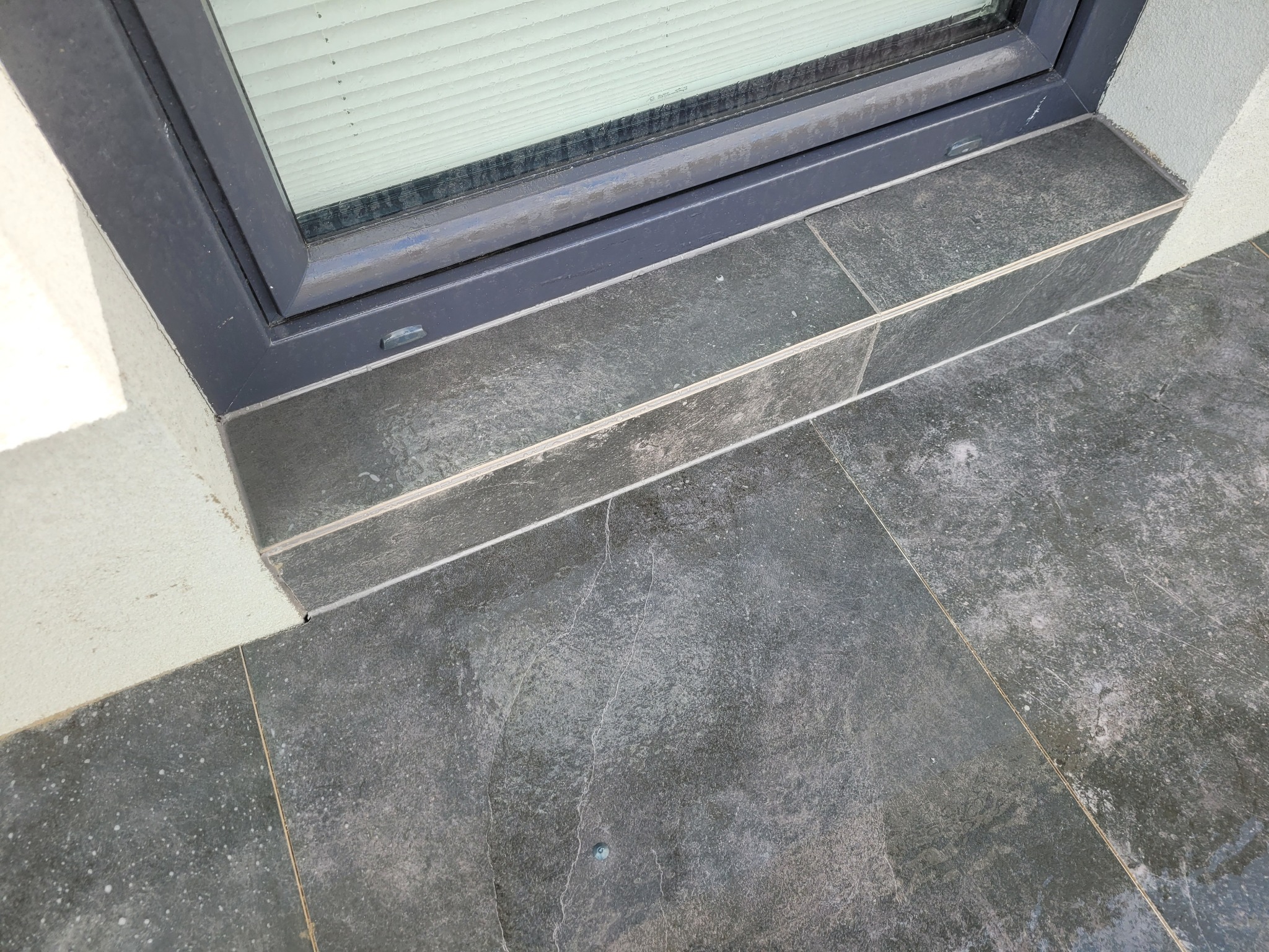 Szare, kwadratowe płytki tarasowe ułożone przy oknie z szarą ramą, widoczne fugi i struktura płytek.