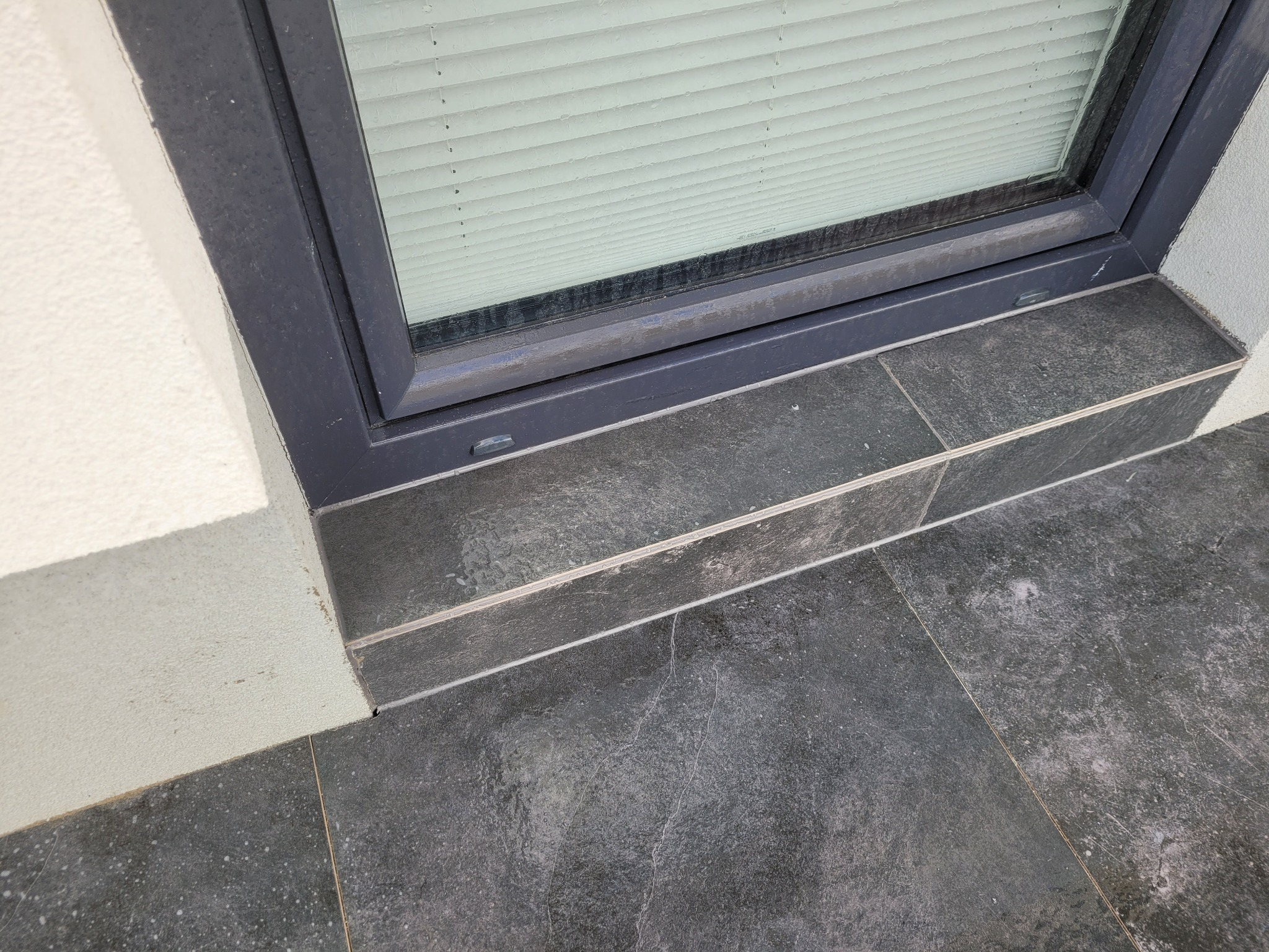 Szare płytki kamienne ułożone jako parapet zewnętrzny pod oknem z żaluzjami, widoczny fragment białej ściany budynku.