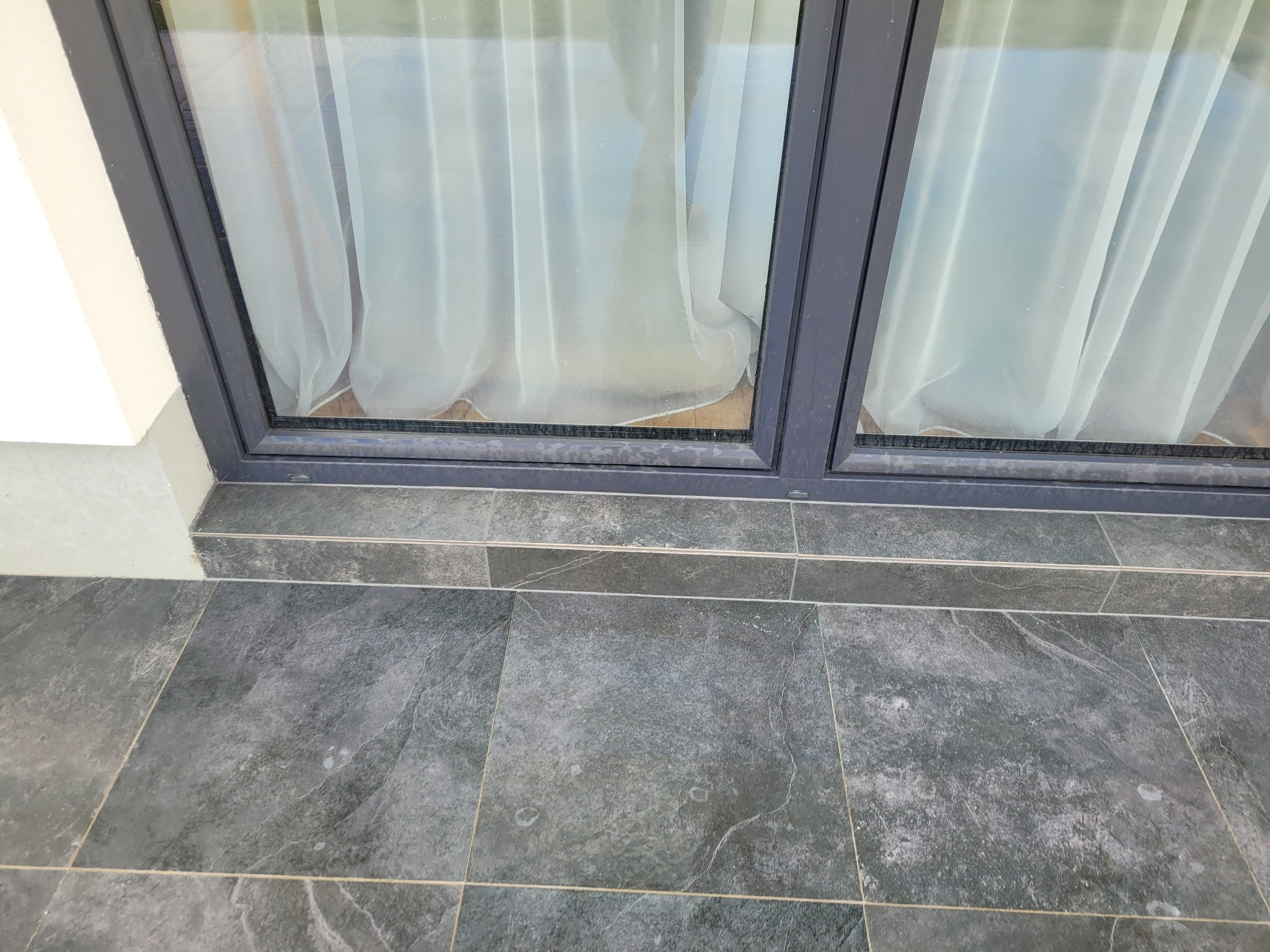 Szare płytki tarasowe ułożone przy oknie z zasłoną, widoczne fugi i fragment ściany.