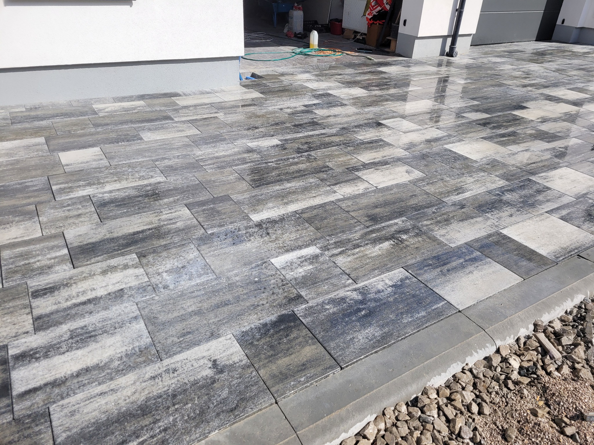 Nowo ułożona, szara kostka brukowa o nieregularnym wzorze, przylegająca do białej ściany budynku z szarym cokołem, obrzeża wykończone szarym krawężnikiem i podsypane drobnym żwirem.