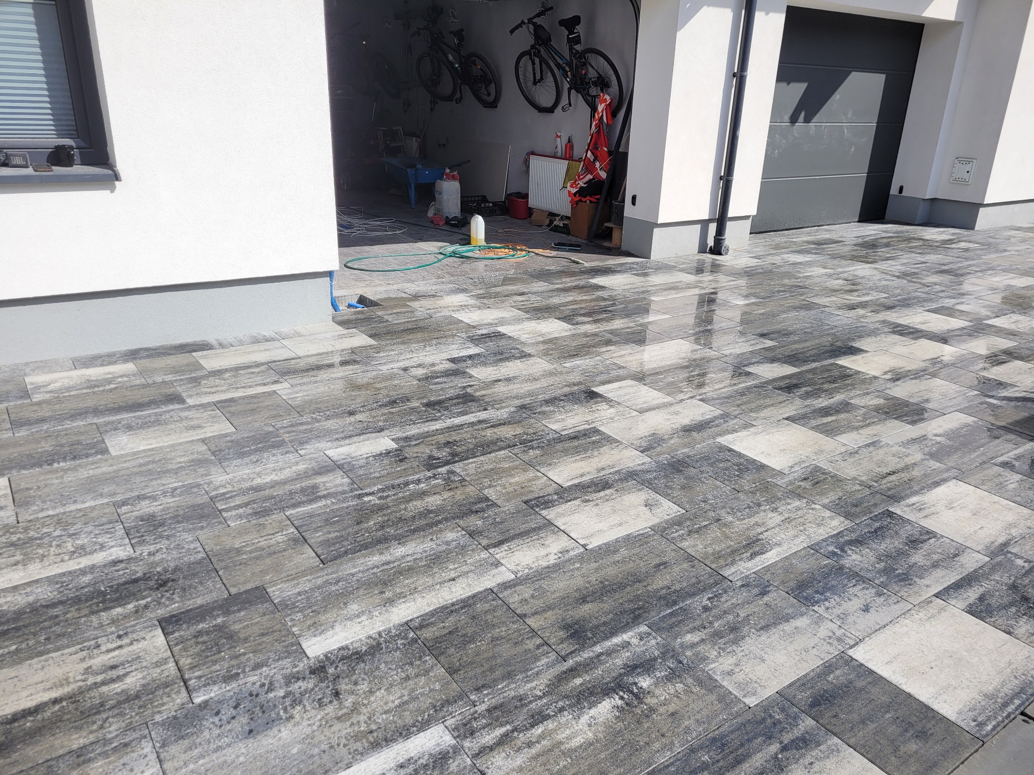 Nowo ułożona, wilgotna kostka brukowa w odcieniach szarości i bieli przed garażem z otwartymi drzwiami, w których widać rowery i narzędzia.