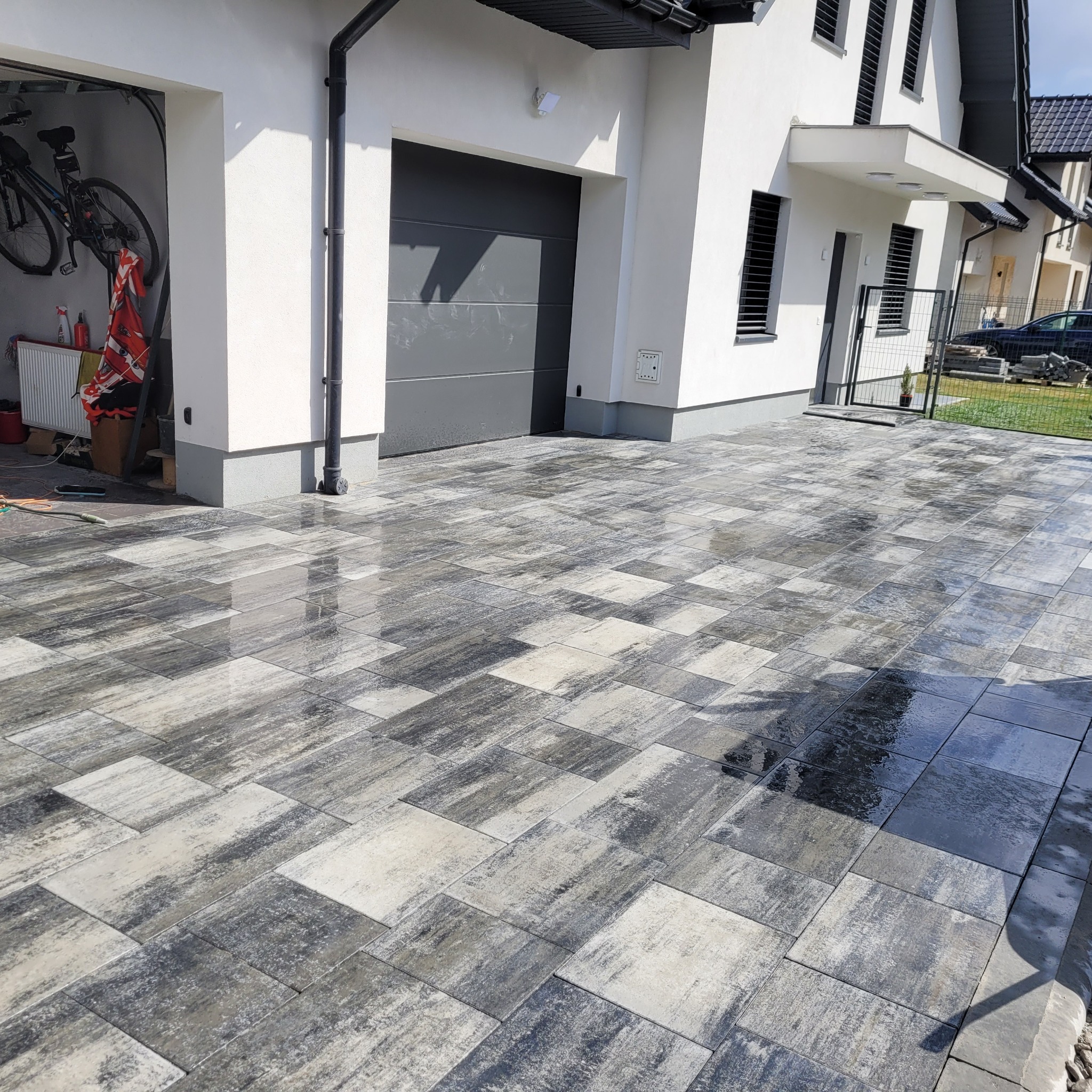 Świeżo ułożona, mokra kostka brukowa w odcieniach szarości i bieli, tworząca wzór na podjeździe przed nowoczesnym domem z szarą bramą garażową i białymi ścianami. W tle widoczny rower w garażu.