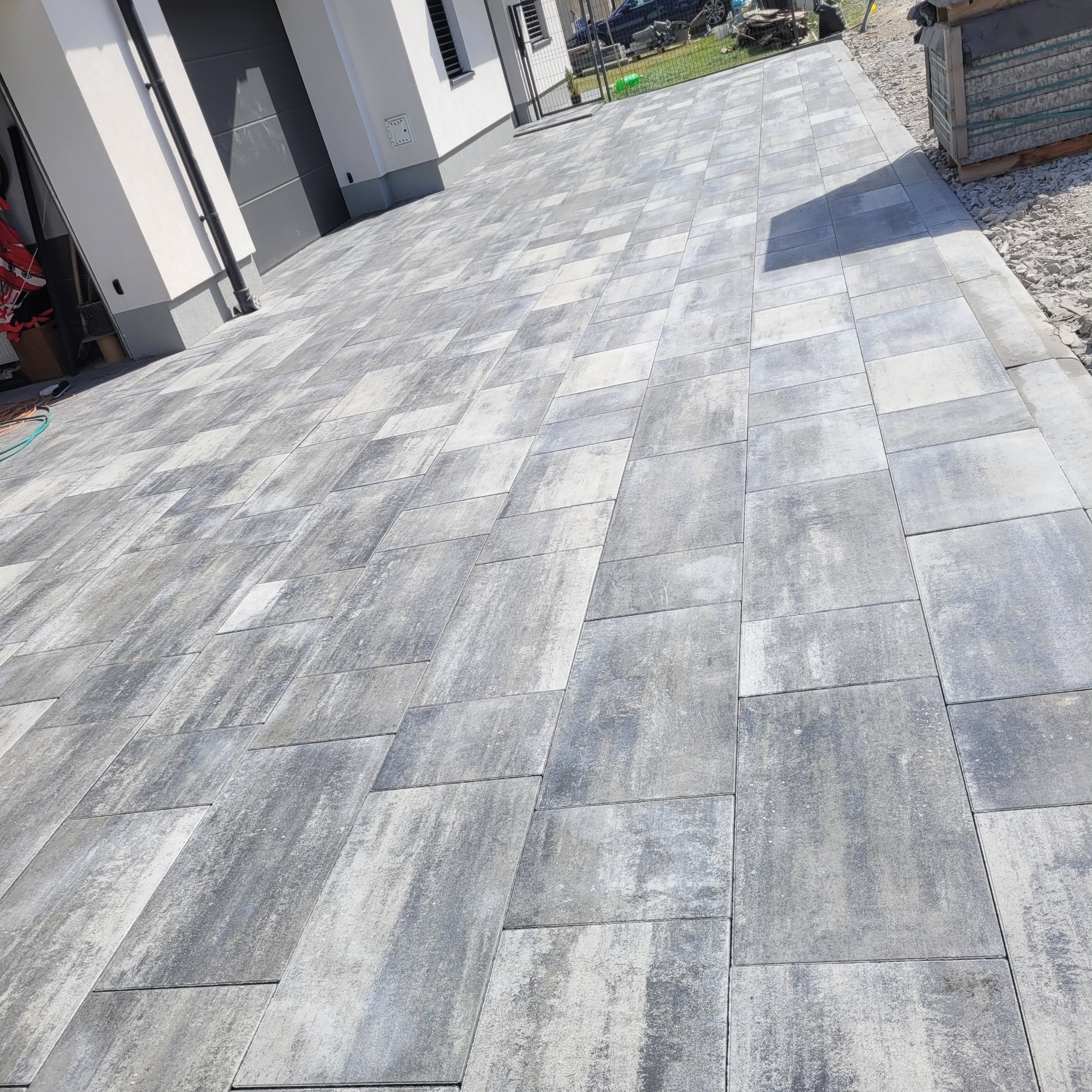 Świeżo ułożona nawierzchnia z szarych, prostokątnych płyt betonowych o zróżnicowanej fakturze, prowadząca do garażu z bramą segmentową i białej elewacji budynku.